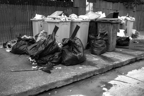 The image shows five large wheelie bins placed on a concrete pavement, with four being light grey and one dark grey, all filled with various types of rubbish. Some bins are overflowing, with waste spilling onto the ground, including black plastic rubbish bags, crumpled paper, and miscellaneous debris. Surrounding the bins, several tightly packed black plastic bags are positioned on the pavement, some leaning against the bins and others scattered nearby. The background features a wooden fence, typical of residential or commercial boundary fencing, with a segment of a garden or outdoor area behind it. The scene suggests a typical waste collection or rubbish disposal setup, with visible clutter and waste materials awaiting removal. The overall environment appears to be an outdoor space associated with a property or business in Limehouse, referencing local rubbish collection practices, with the scene presenting a snapshot of waste awaiting collection by a professional rubbish removal service such as Office Clearance Limehouse. The lighting is even, indicating a daytime setting, highlighting the textures of the plastic bags, the rough concrete, and the smooth surfaces of the bins.