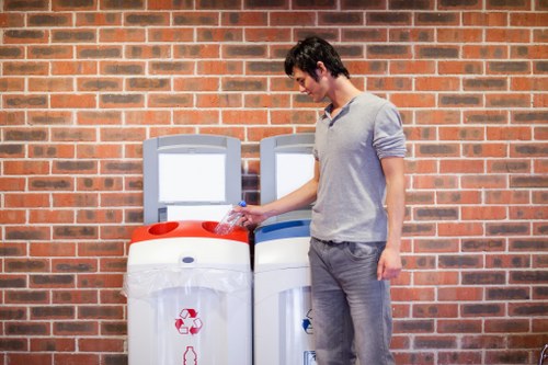 A young man with dark hair, wearing a light grey polo shirt and grey trousers, is seen disposing of a plastic water bottle into a white recycling bin with a red recycling symbol, positioned next to another similar bin. These bins are standing outdoors on a paved area adjacent to a red brick wall, suggesting a commercial or urban environment. The bins have a smooth, plastic surface with a small slot at the top for placing recyclables. The background features a brick wall with visible mortar joints, and the lighting appears natural, illuminating the scene evenly. This image illustrates rubbish disposal or recycling practices in an urban setting, relevant to rubbish removal services provided by Office Clearance Limehouse, located in the London area, promoting environmentally responsible waste management.