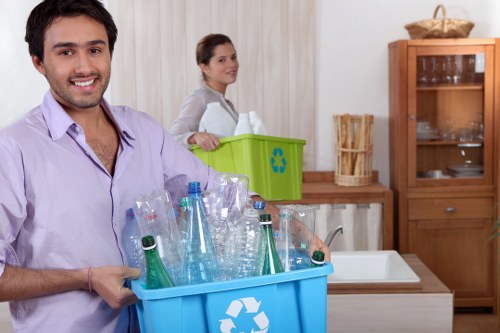 A man wearing a light purple shirt is smiling and holding a large blue recycling bin filled with numerous clear plastic bottles, some with green and clear caps, mixed with other plastic waste. Behind him, a woman dressed in a light-colored top is standing in a kitchen area, next to a green recycling box with a blue recycling symbol, placed on a wooden countertop. The background includes a wooden cabinet with shelves and glass doors, containing various glassware, and a wicker basket on top. The kitchen has neutral-toned curtains and a modern, clean appearance. The scene depicts an indoor setting focused on waste collection and recycling, consistent with rubbish removal services such as those offered by Office Clearance Limehouse, facilitating environmentally responsible disposal of waste material in the local area near Limehouse or the London E14 postcode, in a professional and factual manner.