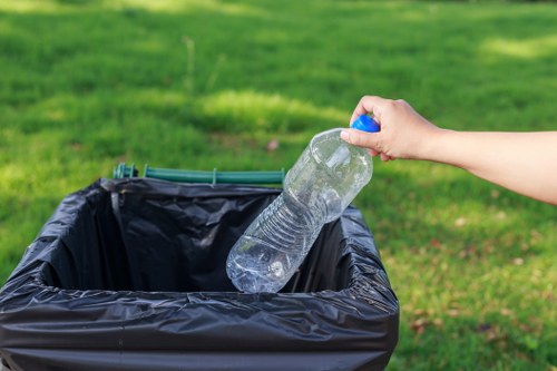A person’s hand is shown holding a clear plastic water bottle with a blue cap, about to drop it into an open black rubbish bin lined with a black plastic bag. The background features a green grassy area, suggesting the scene is outdoors in a park or garden setting. The environment appears well-lit with natural daylight, and the focus is on the act of disposal of recyclable plastic waste, aligned with rubbish removal services like those offered by Office Clearance Limehouse. The scene emphasizes responsible waste disposal and environmental cleanliness, situated in a typical outdoor space near residential or commercial property in Limehouse or nearby London areas, subtly supporting local rubbish collection and recycling efforts.