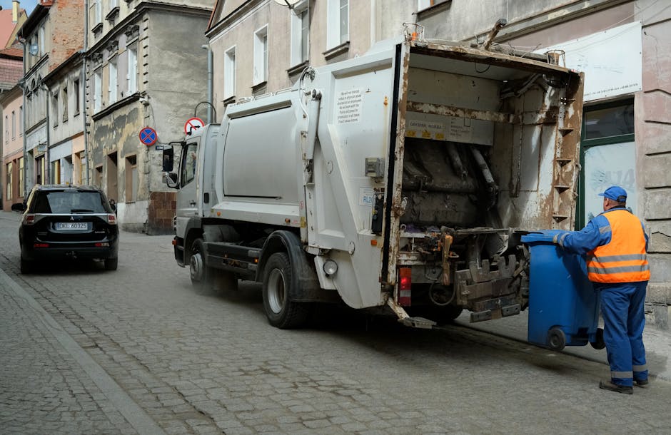 A worker stands beside a large red garbage truck on a roadside, loading waste into the rear compactor section. The worker is wearing a fluorescent yellow and orange safety vest over dark clothing and 