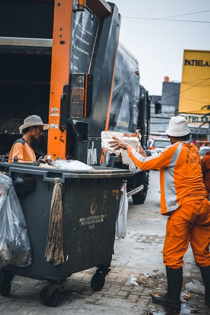 A man wearing a face mask, grey shirt, and black gloves is standing on the edge of a large, open-top black waste collection truck parked outdoors in an urban environment. He is lifting or sorting thro