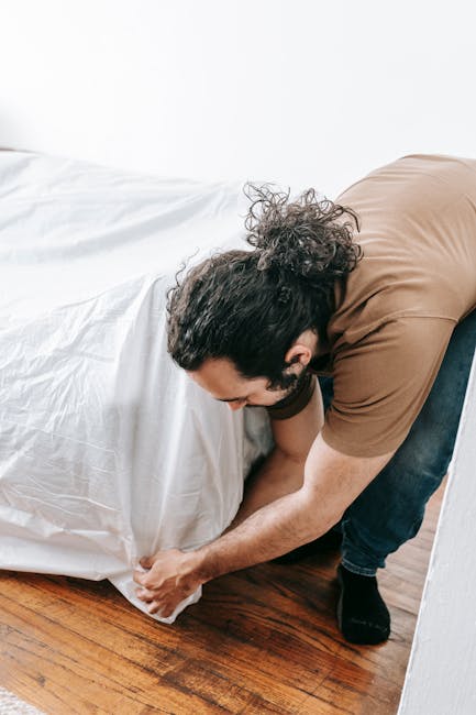 A man with dark, curly hair tied back in a bun, wearing a brown t-shirt and dark jeans, is crouched down on a wooden floor, lifting the corner of a large white plastic rubbish bag. The bag appears to