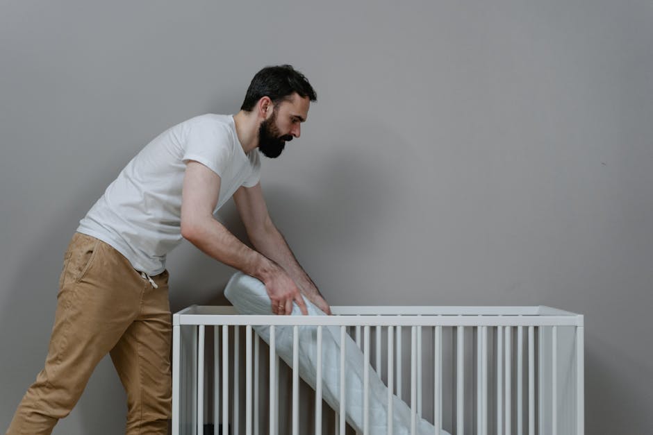 A man with dark, curly hair tied back in a bun, wearing a brown t-shirt and dark jeans, is crouched down on a wooden floor, lifting the corner of a large white plastic rubbish bag. The bag appears to