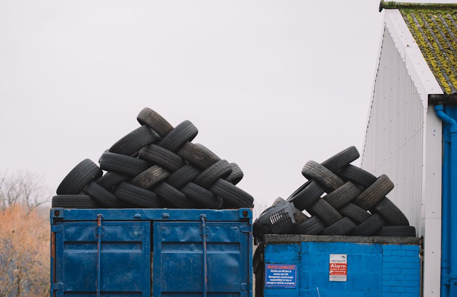 An open landfill site showing a large area covered in mixed waste, including plastic bags, cardboard, and other household refuse, with numerous people actively sorting and collecting trash. Two yellow