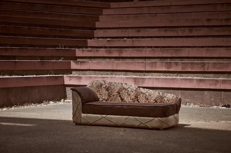 An old, worn-out sofa with a floral patterned fabric on the backrest and armrest, and a dark brown velvet material on the seat cushion, is situated outdoors on a paved surface. The sofa shows signs of